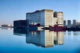 A large industrial building is reflected in calm water, with a red ship moored alongside it. The building has a worn, weathered appearance, and several high-rise residential buildings are visible in the background. The sky is clear and blue, enhancing the serene ambiance of the scene.