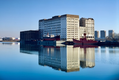 A large industrial building is reflected in calm water, with a red ship moored alongside it. The building has a worn, weathered appearance, and several high-rise residential buildings are visible in the background. The sky is clear and blue, enhancing the serene ambiance of the scene.