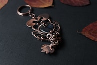 An intricately designed metal keychain lies on a dark textured surface. The keychain features elaborate wirework resembling a fish, with a central dark blue or black stone accent. Surrounding it are a few dried reddish-brown leaves, adding a rustic touch.