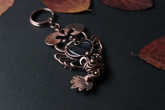An intricately designed metal keychain lies on a dark textured surface. The keychain features elaborate wirework resembling a fish, with a central dark blue or black stone accent. Surrounding it are a few dried reddish-brown leaves, adding a rustic touch.