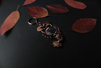 A decorative brass keychain with intricate designs lies on a dark surface, surrounded by several dried, reddish-brown leaves. The keychain features a central blue stone and elaborate metalwork resembling an ornamental figure.