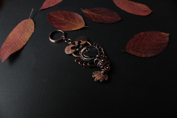 A decorative brass keychain with intricate designs lies on a dark surface, surrounded by several dried, reddish-brown leaves. The keychain features a central blue stone and elaborate metalwork resembling an ornamental figure.