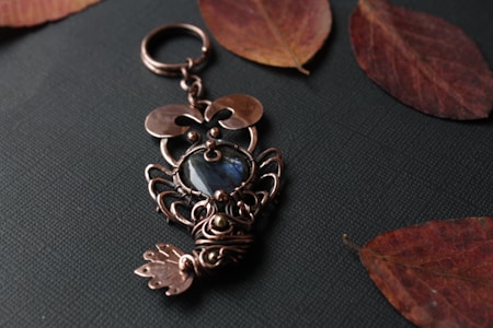 An ornate metal keychain with a design resembling an owl, featuring a blue gemstone at the center. The keychain is placed on a dark textured surface, surrounded by several dried leaves, creating an autumnal atmosphere.
