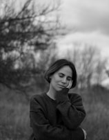 A woman stands in a relaxed posture with her eyes closed and one hand resting on her cheek. She wears a dark sweater, and the background shows blurred trees and cloudy skies, giving a serene outdoor setting.