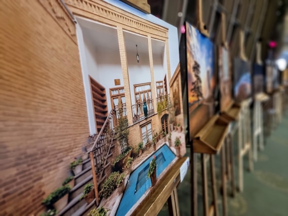 A collection of framed photos displayed on easels in an art gallery setting. The focus is on a photograph showing a traditional architectural structure with a small courtyard, featuring tall pillars, wooden doors, and a blue-tiled pool surrounded by plants. Other photos are displayed in the background, slightly out of focus.