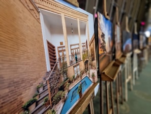 A collection of framed photos displayed on easels in an art gallery setting. The focus is on a photograph showing a traditional architectural structure with a small courtyard, featuring tall pillars, wooden doors, and a blue-tiled pool surrounded by plants. Other photos are displayed in the background, slightly out of focus.
