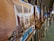 A collection of framed photos displayed on easels in an art gallery setting. The focus is on a photograph showing a traditional architectural structure with a small courtyard, featuring tall pillars, wooden doors, and a blue-tiled pool surrounded by plants. Other photos are displayed in the background, slightly out of focus.