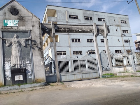 A large concrete building with several stories and multiple windows features prominently in the image. The foreground displays tall metal gates, and a statue of a woman with outstretched arms holding scales is mounted on the wall, suggesting a legal or judicial theme. The building and surrounding structures exhibit signs of aging and weathering.