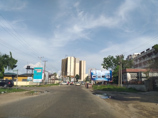 A street scene with a multi-story building in the background. There are billboards on both sides of the road, one advertising 'Breaking News' and another with images of vehicles. Several cars are parked along the street, and there are some trees and electric poles visible. The sky is clear with some clouds.