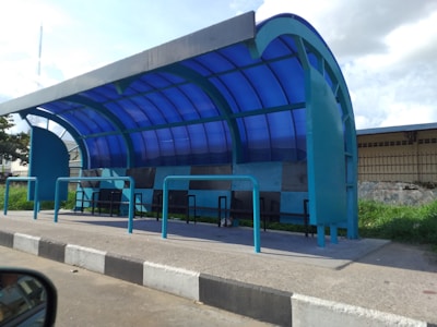 A modern bus shelter with a curved blue-tinted roof and teal-colored framework. The shelter has several black benches underneath the transparent roof, which allows natural light to pass through. It is located beside a road with a concrete sidewalk and black-and-white striped curb. There is greenery behind the shelter, along with a building and some trees.