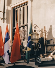 Several national flags hang outside a building, which features decorative architectural elements and signage in multiple languages. The scene is lit by warm sunlight, casting intricate shadows against the wall.