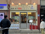 A storefront with a sign that reads 'Yummy, Udon Gyoza' in both English and Japanese. The entrance has a glass door with a menu displayed on the window, showcasing various dishes. Two people are standing outside, observing the store. The exterior includes a mix of brick and beige walls, with a few decorative lights above the sign.