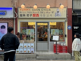 A storefront with a sign that reads 'Yummy, Udon Gyoza' in both English and Japanese. The entrance has a glass door with a menu displayed on the window, showcasing various dishes. Two people are standing outside, observing the store. The exterior includes a mix of brick and beige walls, with a few decorative lights above the sign.