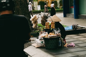 A street vendor with a traditional conical hat is selling various baked goods and snacks displayed in baskets on the sidewalk. The scene includes pedestrians walking by, and the setting appears to be an urban area with trees and buildings in the background.