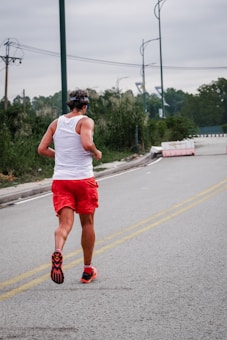 A person is running along a paved road wearing a white tank top, red shorts, and red running shoes. The surroundings include greenery with trees and bushes, and a cloudy sky overhead. Streetlights and barriers are also visible along the road.