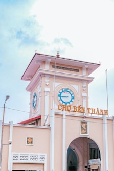 A historic market building with a prominent clock tower and the name 'Chợ Bến Thành' displayed in large letters. The architecture features a blend of colonial and traditional Vietnamese style with decorative elements. The sky is partly cloudy, contributing to a bright and open atmosphere.