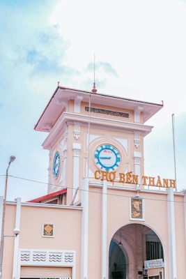 A historic market building with a prominent clock tower and the name 'Chợ Bến Th&agrave;nh' displayed in large letters. The architecture features a blend of colonial and traditional Vietnamese style with decorative elements. The sky is partly cloudy, contributing to a bright and open atmosphere.