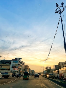The image captures a bustling urban street scene during sunset. Several vehicles, including a tuk-tuk and cars, are seen driving along the road. People are walking on the sidewalks, and birds are flying in the sky. Buildings with advertisements line the street, and there are power lines with birds perched along them.