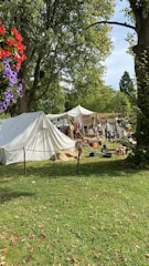 A historical reenactment scene with tents set up in a grassy area surrounded by tall trees. People in period clothing are engaged in various activities. Some red and purple flowers are visible in the foreground.