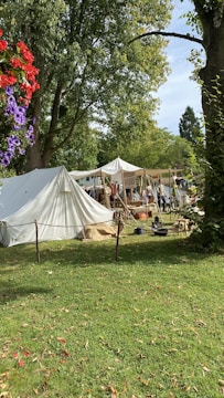 A historical reenactment scene with tents set up in a grassy area surrounded by tall trees. People in period clothing are engaged in various activities. Some red and purple flowers are visible in the foreground.