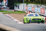 A vibrant green race car, specifically a Mercedes-AMG GT3, is speeding on a racetrack. The track features red and white barriers, and the background includes trees and a digitally displayed screen showing various graphics. Pit crew members in orange outfits are visible off to the side of the track.