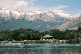 People enjoy various water activities on a calm lake surrounded by lush greenery. A pavilion sits near the water's edge with snow-capped mountains in the background under a partly cloudy sky.