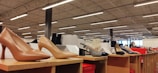 Various styles of women's high-heeled shoes are displayed on wooden shelves inside a well-lit store. Price tags can be seen near some of the shoes, and the ceiling features exposed beams and fluorescent lighting.