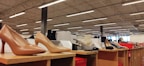 Various styles of women's high-heeled shoes are displayed on wooden shelves inside a well-lit store. Price tags can be seen near some of the shoes, and the ceiling features exposed beams and fluorescent lighting.