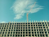 A high-rise building under construction with a tower crane beside it, set against a clear blue sky with some clouds. The building structure is prominently displayed with multiple floors and exposed concrete pillars.