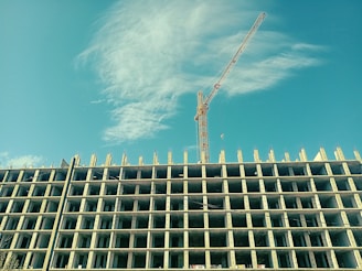 A high-rise building under construction with a tower crane beside it, set against a clear blue sky with some clouds. The building structure is prominently displayed with multiple floors and exposed concrete pillars.