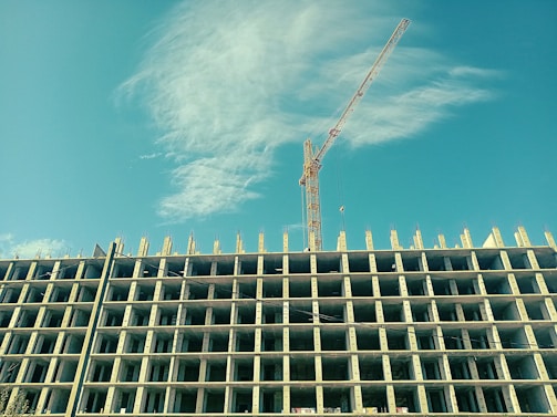 A high-rise building under construction with a tower crane beside it, set against a clear blue sky with some clouds. The building structure is prominently displayed with multiple floors and exposed concrete pillars.