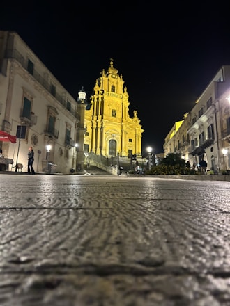 A beautifully illuminated baroque cathedral stands majestically against the night sky, surrounded by historic buildings. The intricate architecture is highlighted by warm yellow lighting, creating a striking contrast with the darkness. Cobblestone pavement leads up to the cathedral, adding to the old-world charm. A few people are visible walking nearby, adding a sense of scale and liveliness.