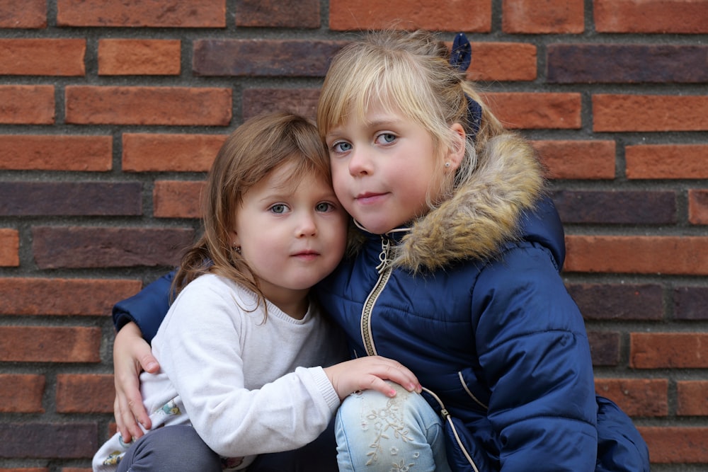 two little girls hugging each other in front of a brick wall