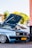 A light blue Volkswagen car with its hood open, showcasing the engine. It is parked in an urban setting with palm trees in the background. The car has customized alloy wheels and a yellow license plate.