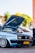 A light blue Volkswagen car with its hood open, showcasing the engine. It is parked in an urban setting with palm trees in the background. The car has customized alloy wheels and a yellow license plate.