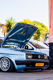 A light blue Volkswagen car with its hood open, showcasing the engine. It is parked in an urban setting with palm trees in the background. The car has customized alloy wheels and a yellow license plate.