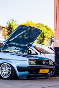 A light blue Volkswagen car with its hood open, showcasing the engine. It is parked in an urban setting with palm trees in the background. The car has customized alloy wheels and a yellow license plate.