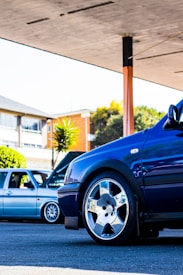 A close-up view of a blue car with shiny, chrome wheels in a parking area. In the background, a light grey car with its hood open is visible. A building with large windows and some greenery, including a large plant, can be seen in the background.