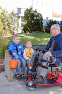 A man on a mobility scooter interacting with two children seated on a bench, all appearing to be having a joyous conversation. The children hold orange buckets, suggesting they might be collecting treats or participating in an event. The backdrop includes bushes and a building, hinting at a park or urban setting.