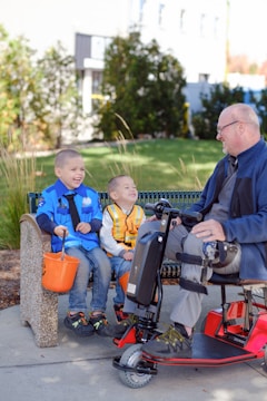 A man on a mobility scooter interacting with two children seated on a bench, all appearing to be having a joyous conversation. The children hold orange buckets, suggesting they might be collecting treats or participating in an event. The backdrop includes bushes and a building, hinting at a park or urban setting.