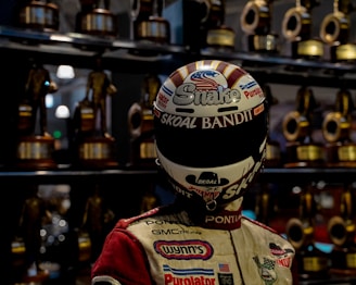 A racing driver wearing a helmet and racing suit stands in front of a display of numerous trophies. The helmet has various logos and text, including 'Skoal Bandit'. The background is filled with blurred trophies, suggesting achievements in racing.