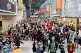 A bustling convention center filled with a large crowd of people, many wearing costumes and cosplay outfits. Banners and signs for the Javits Center are visible, along with promotional displays for various events. Attendees are diverse in age and attire, engaging in conversations and activities typical of a large fan convention.