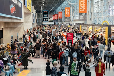 A bustling convention center filled with a large crowd of people, many wearing costumes and cosplay outfits. Banners and signs for the Javits Center are visible, along with promotional displays for various events. Attendees are diverse in age and attire, engaging in conversations and activities typical of a large fan convention.