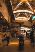 An indoor marketplace features a high, vaulted ceiling with bright windows casting warm light below. People are walking and shopping, with stalls and goods lined along the sides. A motorcycle is parked in the center, and various items can be seen on shelves, adding to the busy atmosphere.