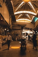 An indoor marketplace features a high, vaulted ceiling with bright windows casting warm light below. People are walking and shopping, with stalls and goods lined along the sides. A motorcycle is parked in the center, and various items can be seen on shelves, adding to the busy atmosphere.