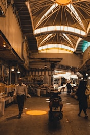 An indoor marketplace features a high, vaulted ceiling with bright windows casting warm light below. People are walking and shopping, with stalls and goods lined along the sides. A motorcycle is parked in the center, and various items can be seen on shelves, adding to the busy atmosphere.