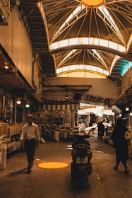 An indoor marketplace features a high, vaulted ceiling with bright windows casting warm light below. People are walking and shopping, with stalls and goods lined along the sides. A motorcycle is parked in the center, and various items can be seen on shelves, adding to the busy atmosphere.
