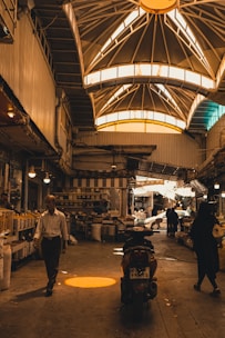 An indoor marketplace features a high, vaulted ceiling with bright windows casting warm light below. People are walking and shopping, with stalls and goods lined along the sides. A motorcycle is parked in the center, and various items can be seen on shelves, adding to the busy atmosphere.