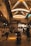 An indoor marketplace features a high, vaulted ceiling with bright windows casting warm light below. People are walking and shopping, with stalls and goods lined along the sides. A motorcycle is parked in the center, and various items can be seen on shelves, adding to the busy atmosphere.