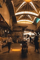 An indoor marketplace features a high, vaulted ceiling with bright windows casting warm light below. People are walking and shopping, with stalls and goods lined along the sides. A motorcycle is parked in the center, and various items can be seen on shelves, adding to the busy atmosphere.